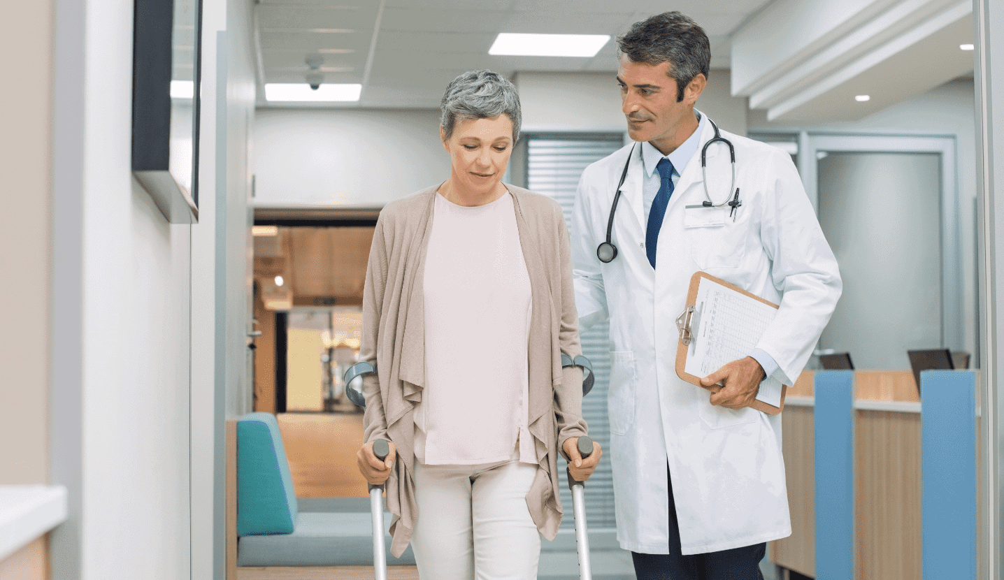 Woman using crutches walks the hall of a hospital with her doctor.