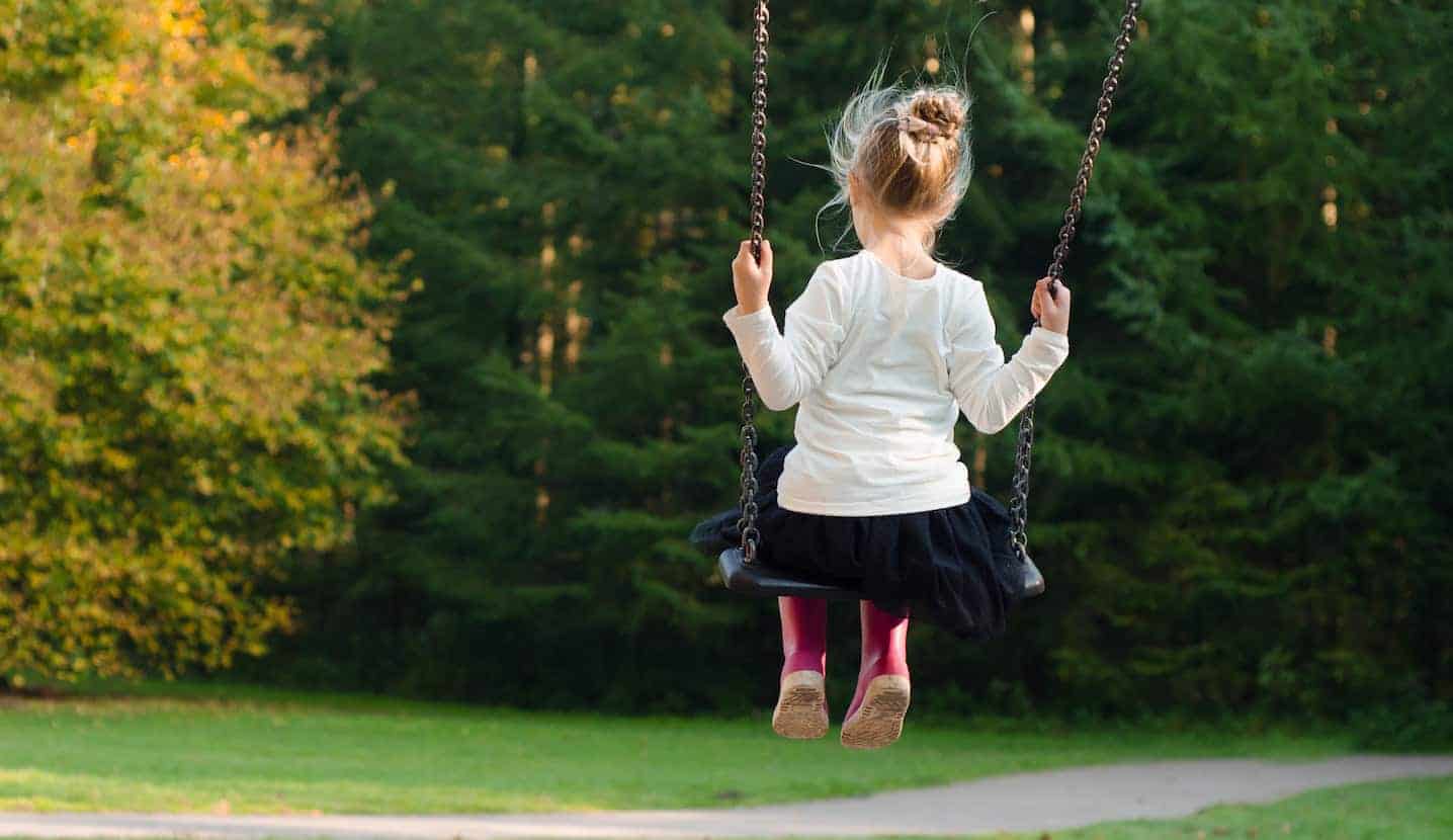 Young girl in a white shirt, black skirt and pink rain boots swinging on a park swing.